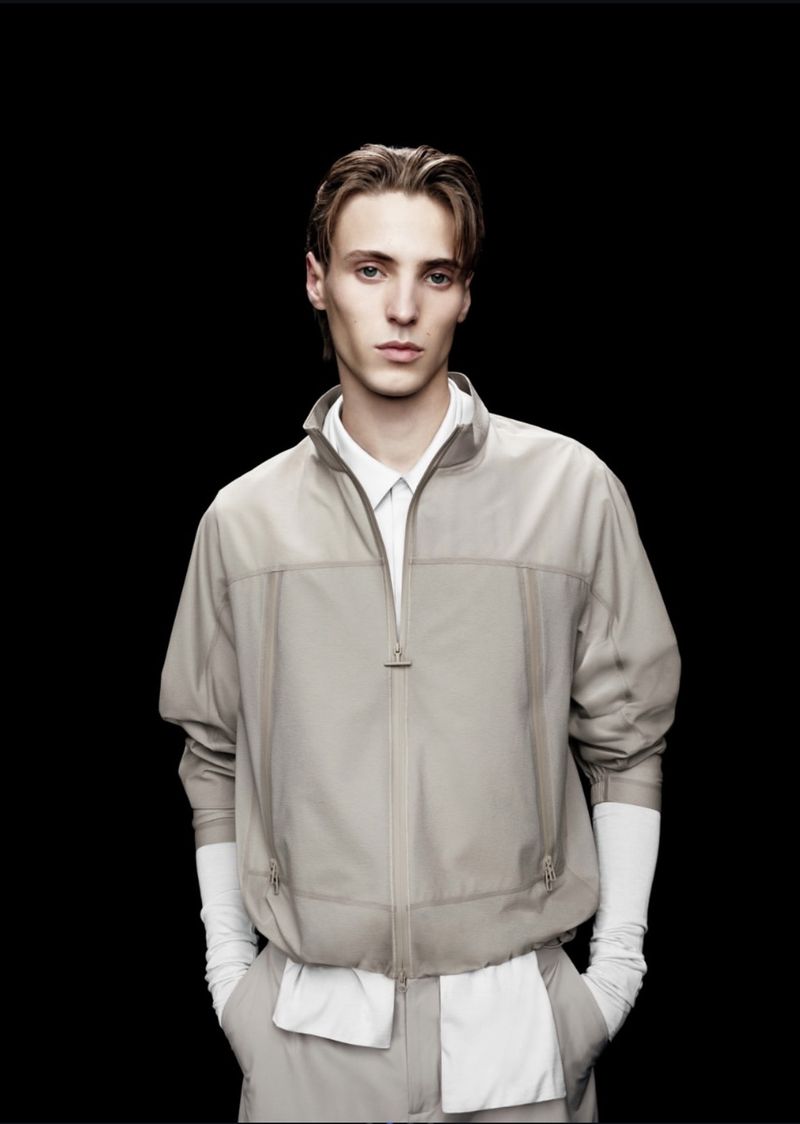 Model Robin Avignon wearing a beige technical zip-up jacket over a white collared shirt against a black seamless backdrop.