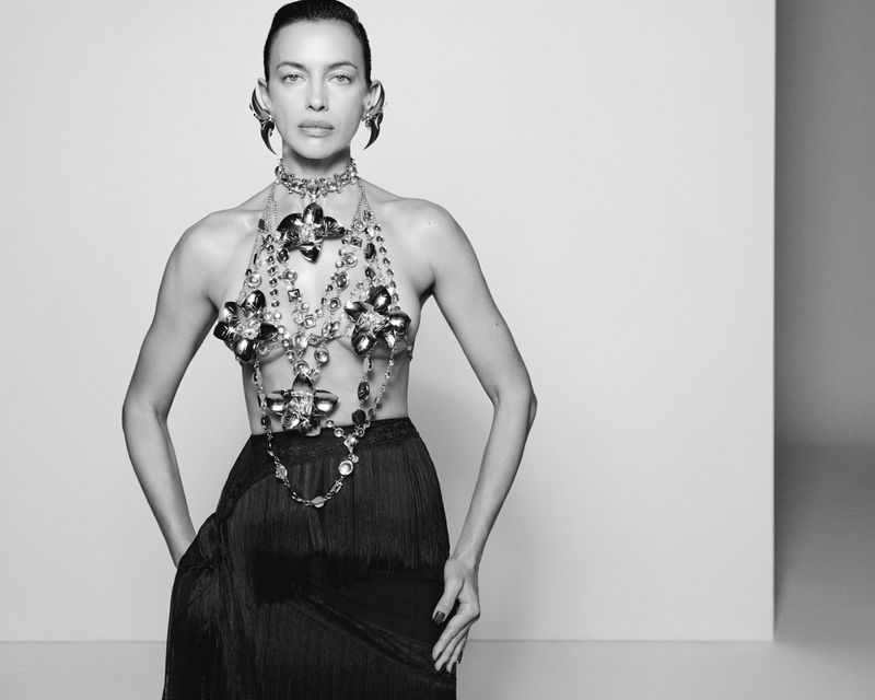Model Irina Shayk wearing a jeweled body harness and fringed skirt against a plain white studio backdrop.