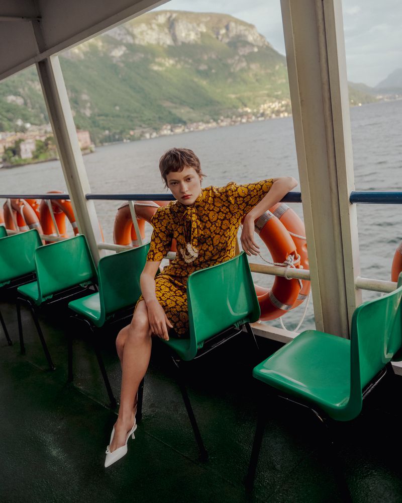 Model wearing a yellow patterned dress seated on a green chair aboard a ferry overlooking a lake and mountains.