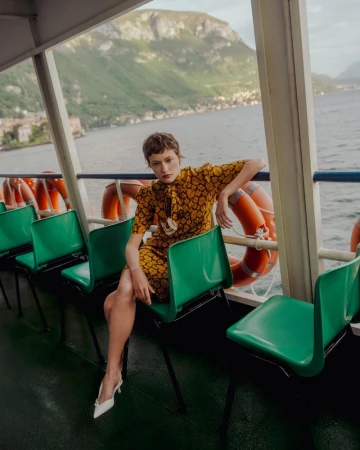Model wearing a yellow patterned dress seated on a green chair aboard a ferry overlooking a lake and mountains.