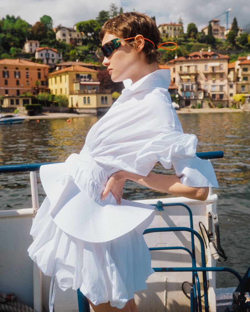Model Daria Zolotova wearing a sculptural white dress and dark sunglasses posing on a boat with hillside Italian buildings behind.