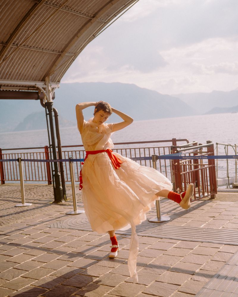 Model Daria Zolotova balancing on one foot in a sheer tiered cream dress with a red sash along a sunlit lakeside promenade.