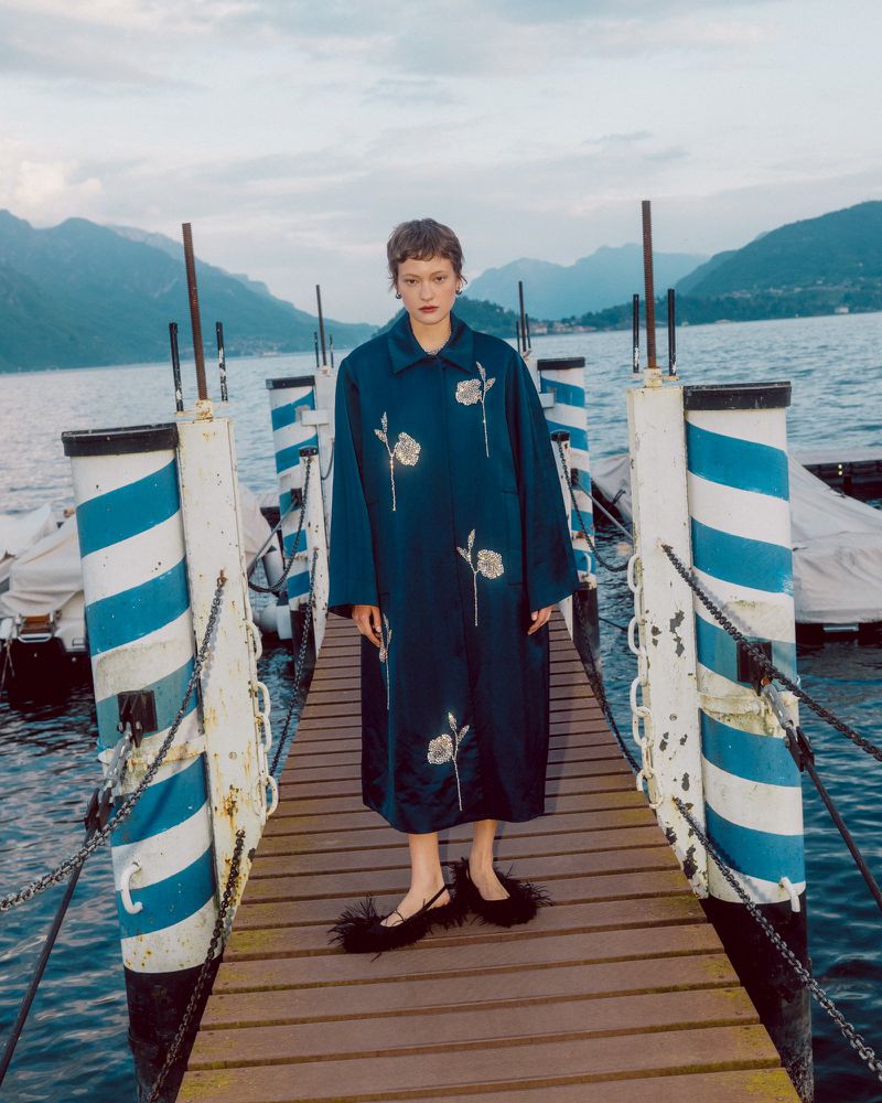 Model standing on a wooden pier wearing a dark teal embroidered coat and feathered shoes with mountains and water behind.