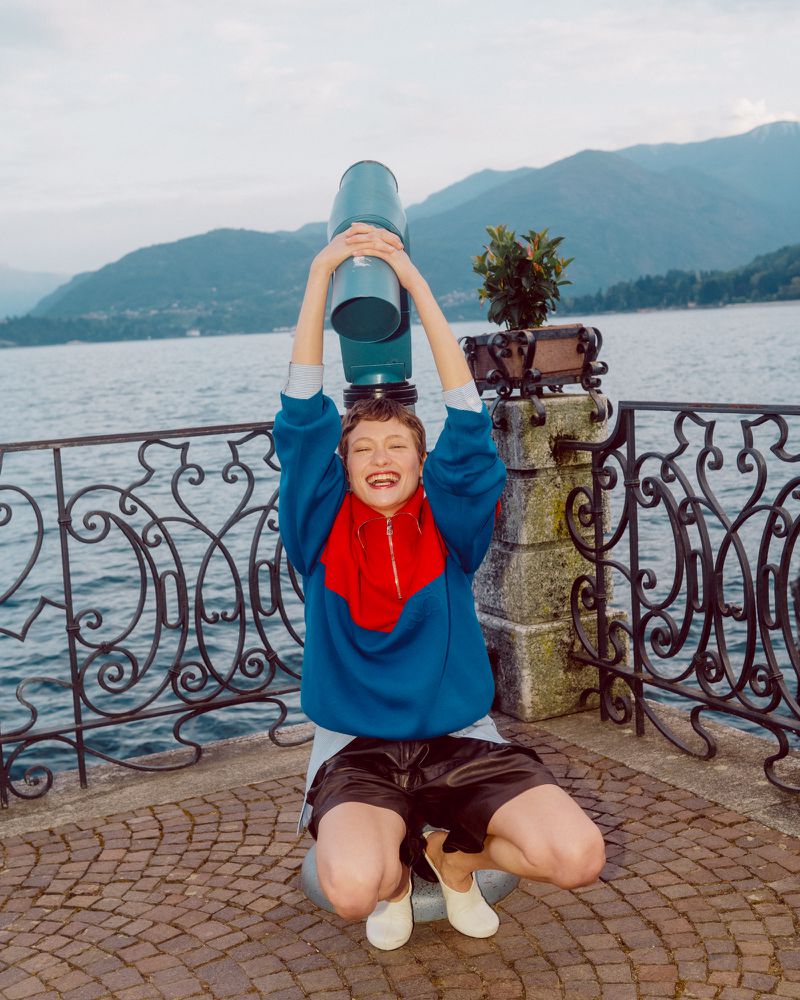 Model Daria Zolotova crouching playfully beneath a viewing telescope along a scenic lakeside promenade.