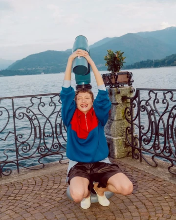Model Daria Zolotova crouching playfully beneath a viewing telescope along a scenic lakeside promenade.