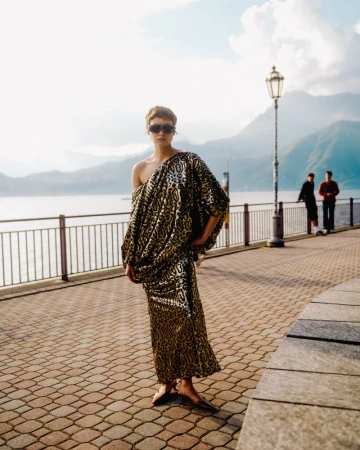 Model Daria Zolotova wearing a metallic draped one-shoulder dress and sunglasses along a scenic lakeside promenade.