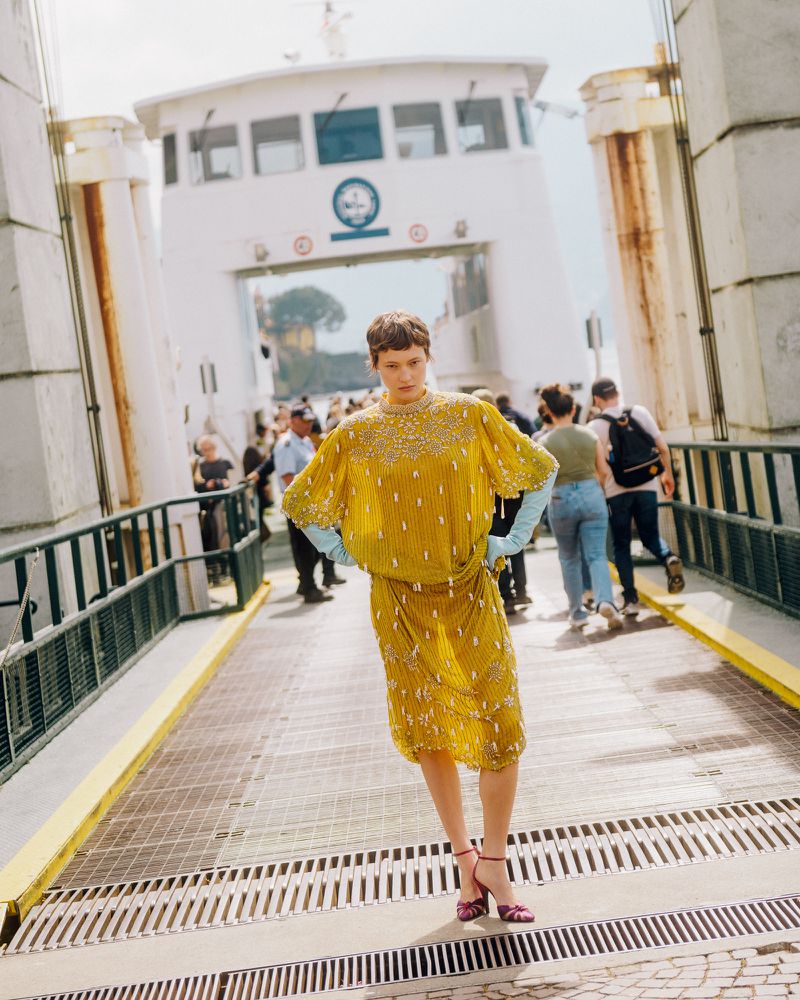 Model Daria Zolotova wearing a yellow beaded midi dress and light blue gloves while standing on a ferry boarding ramp in daylight.