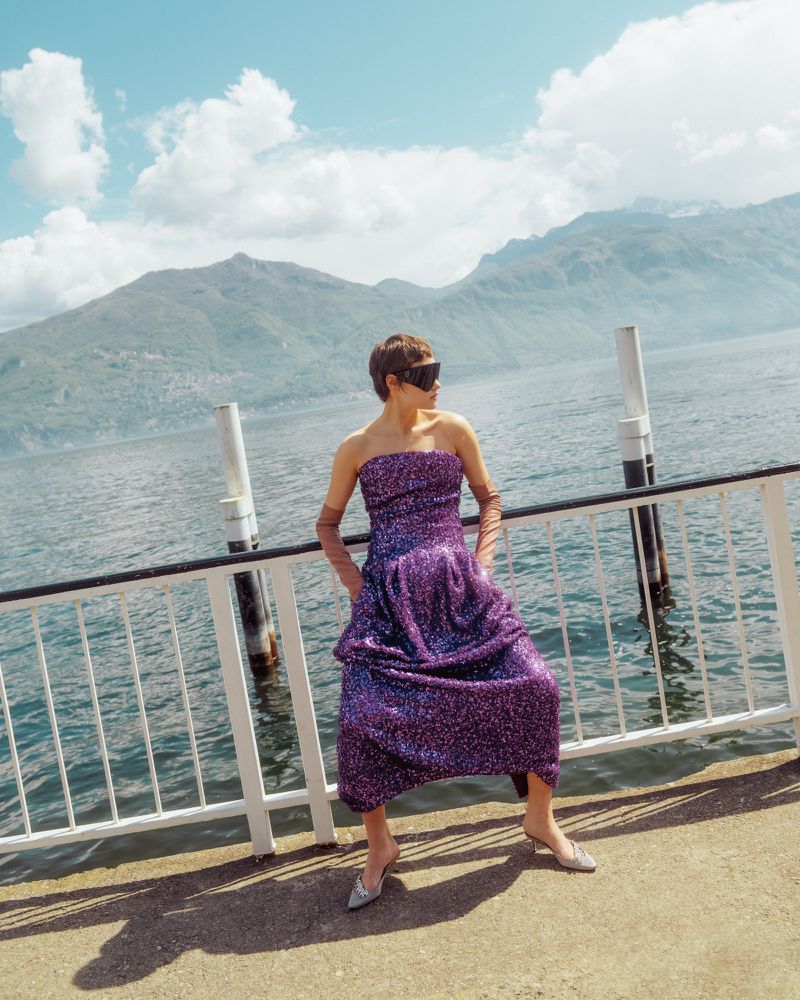 Model Daria Zolotova wearing a strapless purple sequin dress and sunglasses, leaning on a railing by a lake against mountains.