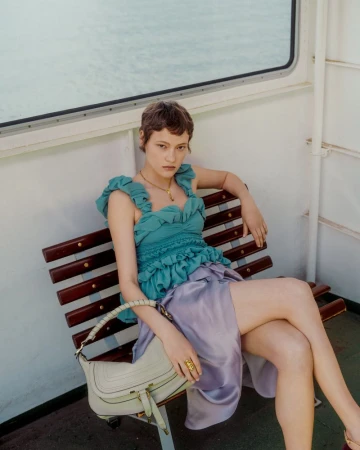 Daria Zolotova seated on a wooden bench in a teal ruffled top and lavender satin skirt with a white leather handbag.