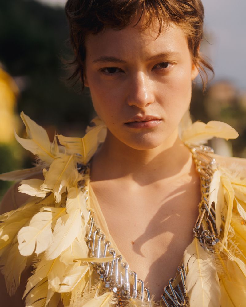 Close-up portrait of a woman with short brown hair wearing a yellow feathered and hardware-embellished top under natural sunlight.