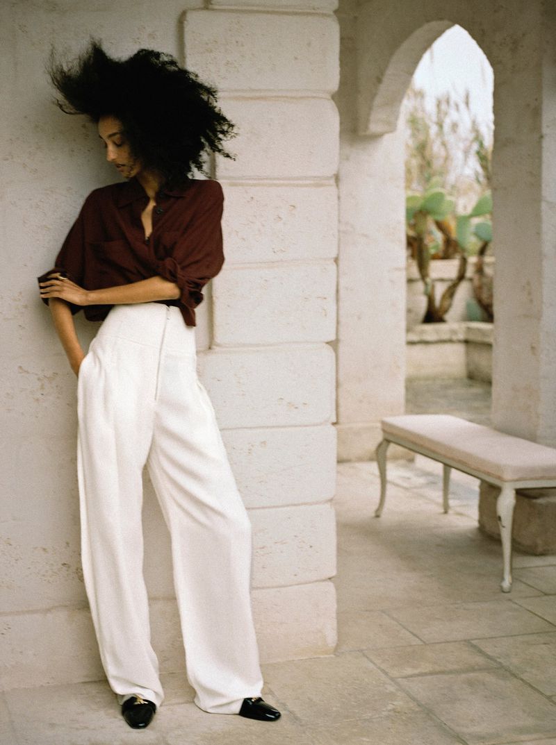 Model Mona Tougaard wearing high-waisted white wide-leg trousers and a brown shirt, leaning against a pale stone archway.