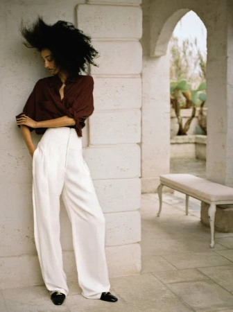 Model Mona Tougaard wearing high-waisted white wide-leg trousers and a brown shirt, leaning against a pale stone archway.