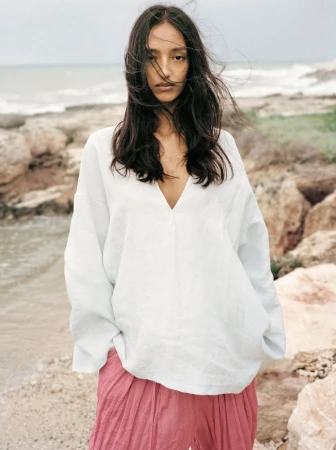 Mona Tougaard wearing an oversized white v-neck tunic and pink trousers on a rocky coastal shoreline.