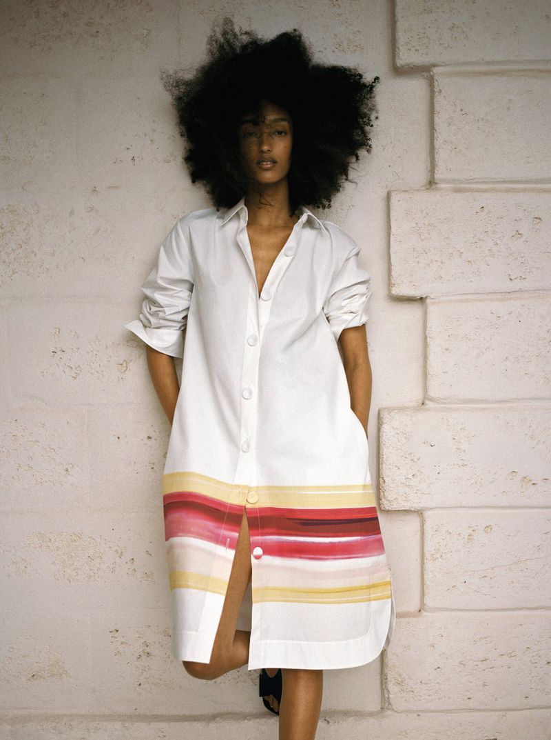 Mona Tougaard wearing a white shirt dress with a colorful striped hem leaning against a textured stone wall.