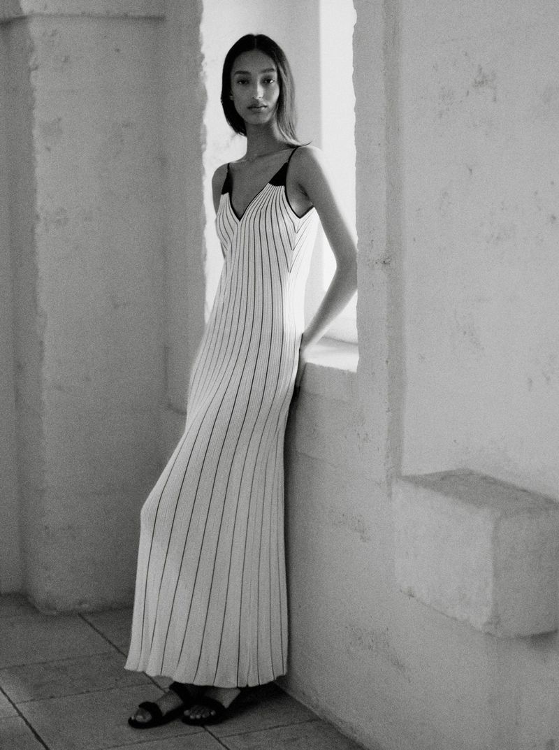 Mona Tougaard in a striped white maxi dress leaning against a wall beside a sunlit window alcove in a black and white portrait.