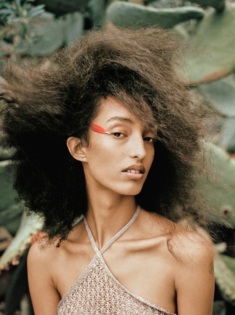 Mona Tougaard with voluminous textured hair and a red feather eye accent posing outdoors in a knit halter top.