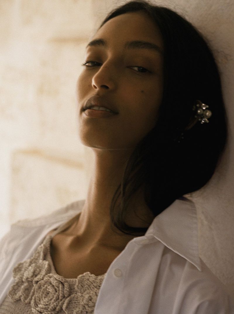 Portrait of an adult woman with long dark hair, wearing a floral top and white collared shirt, leaning against a neutral-toned wall.