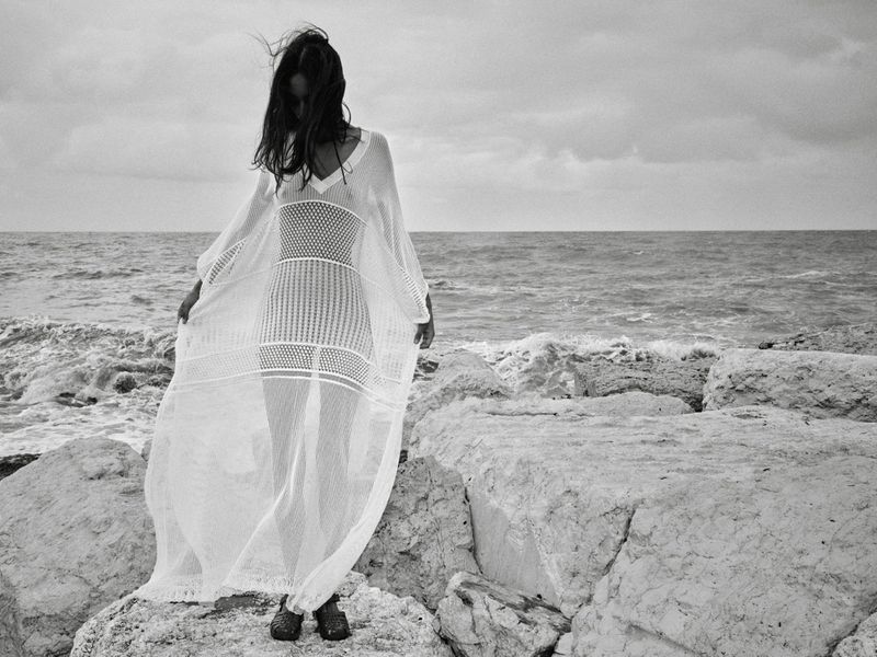Mona Tougaard standing on coastal rocks in a sheer white open-knit caftan dress by the ocean in black and white.