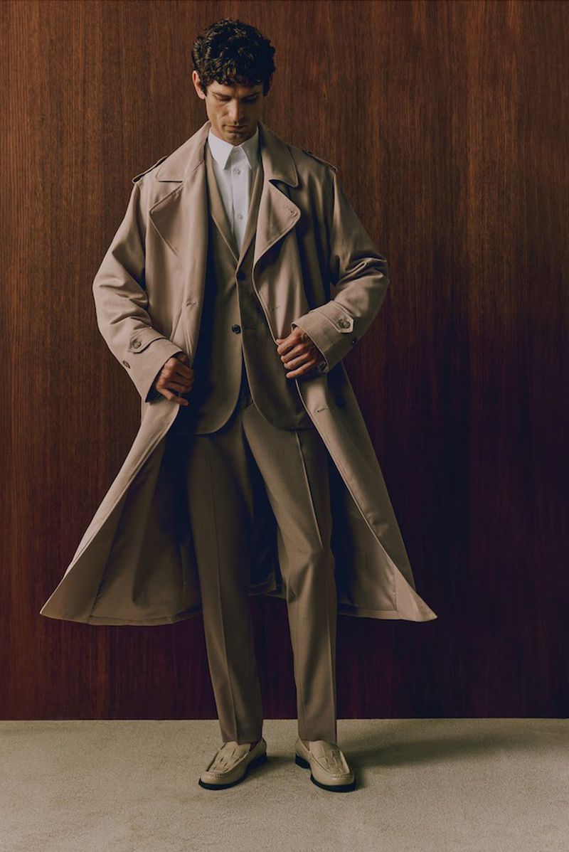 A stylish man in a beige suit, white shirt, and trench coat poses indoors with a wood wall background in a fashion-editorial style.