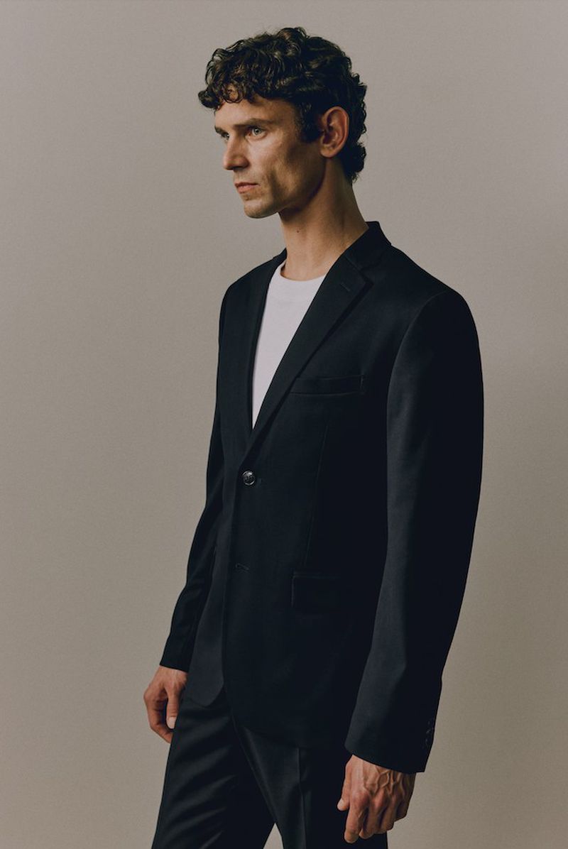 Man in a dark suit and white t-shirt, standing in a photo studio. He is facing left, with a neutral expression.