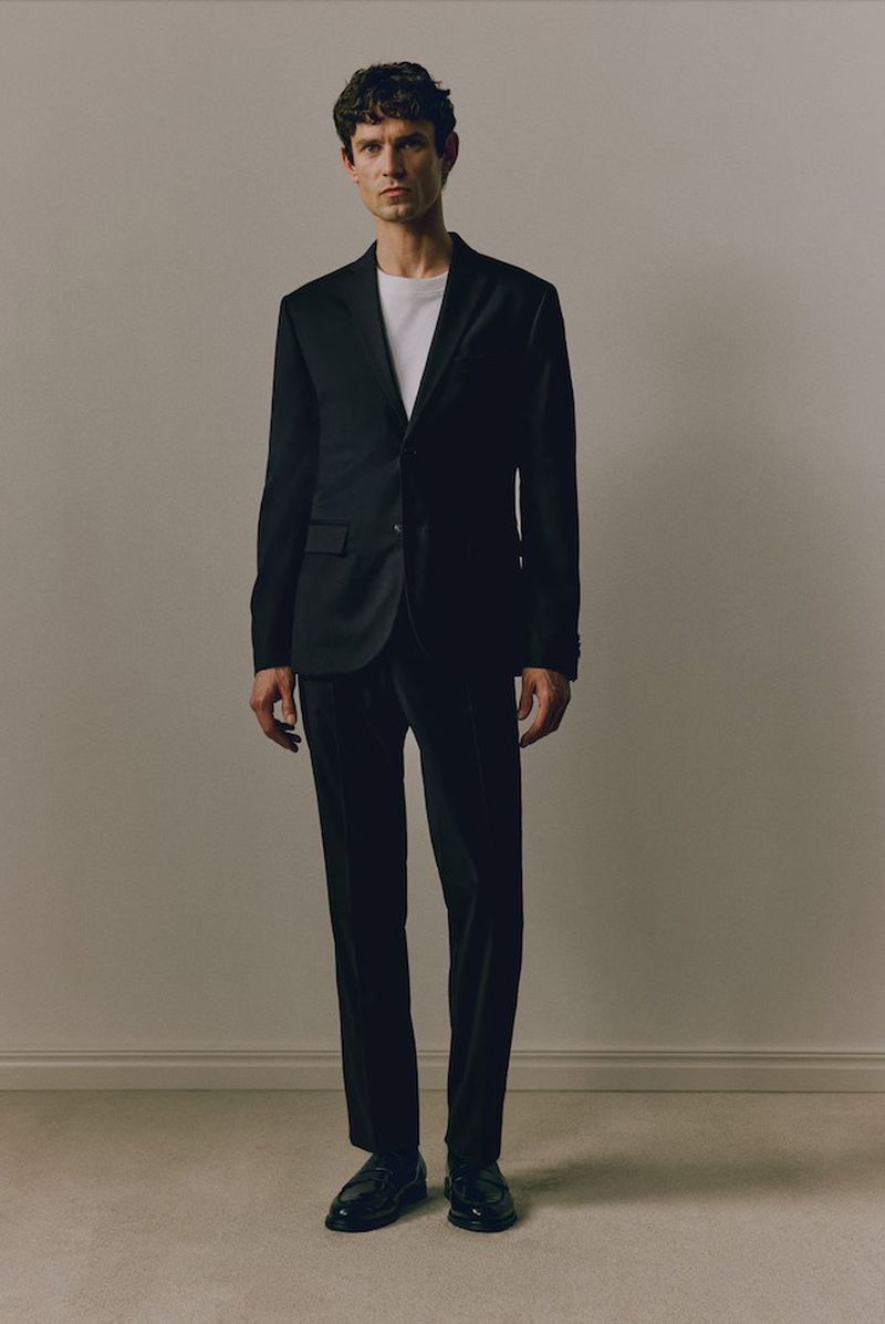 Portrait of a male model in a black suit standing against a neutral background. He is wearing a white shirt underneath the suit.