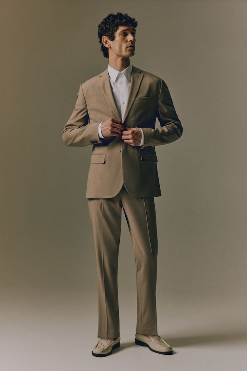 A stylish adult male in a tan suit and white shirt poses in a studio with soft lighting, creating a sophisticated fashion portrait.
