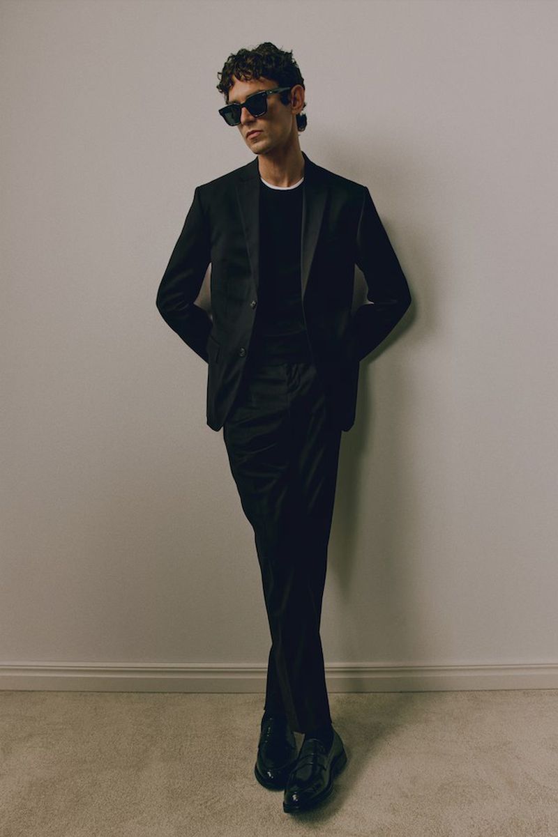 Stylish man in a black suit and sunglasses poses against a neutral wall, showcasing fashion and contemporary menswear design.