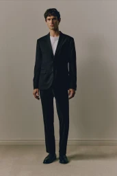 Model Arthur Gosse wearing a black suit and white t-shirt against a neutral grey studio background for H&M lookbook