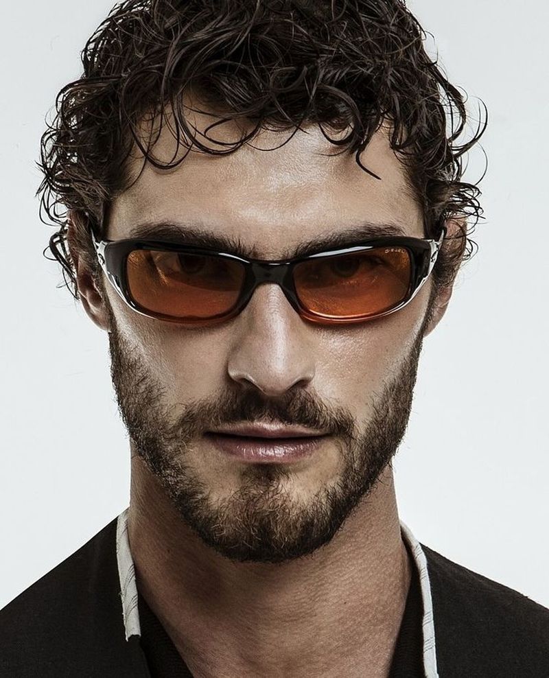 Close-up portrait of model Pedro Pinto wearing tinted sunglasses and a dark collared jacket against a white studio background.