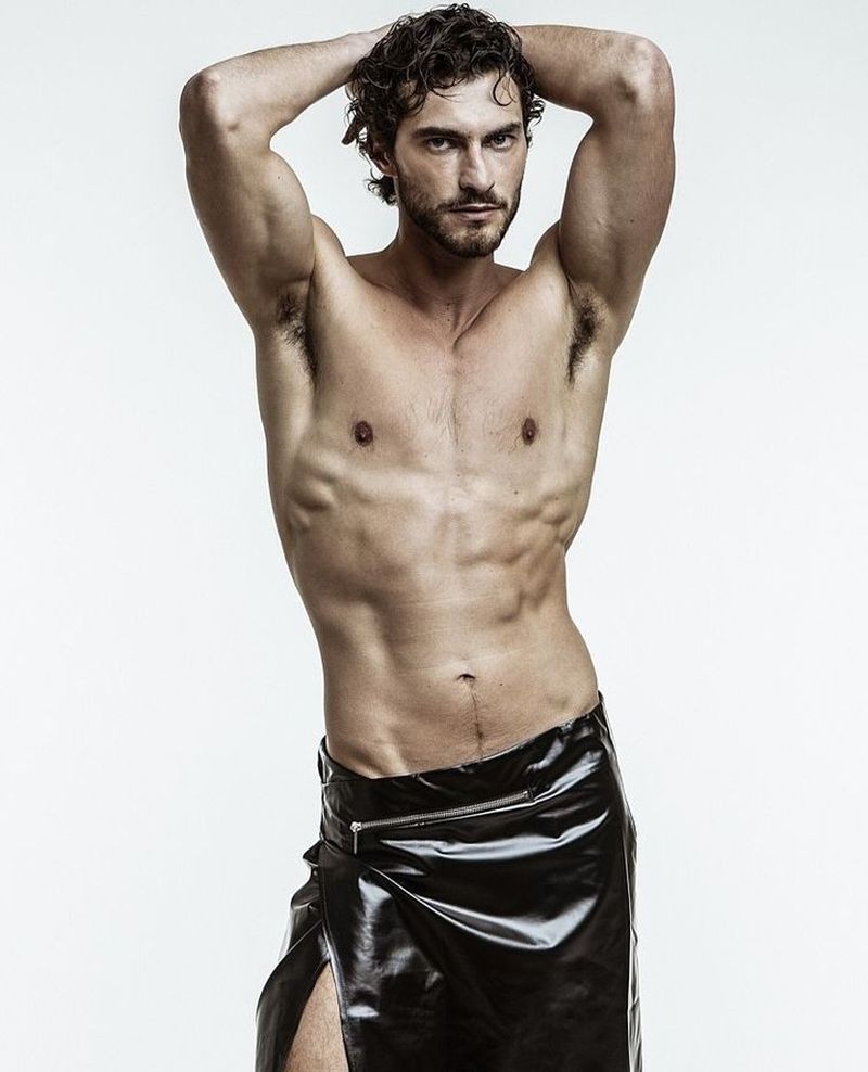 Shirtless man with curly dark hair posing with hands behind his head in a glossy black wrap skirt against a white studio background.
