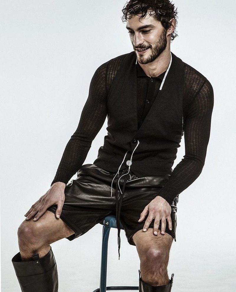 Model Pedro Pinto seated on a stool wearing a black vest, sheer knit polo, leather shorts, and tall black leather boots.