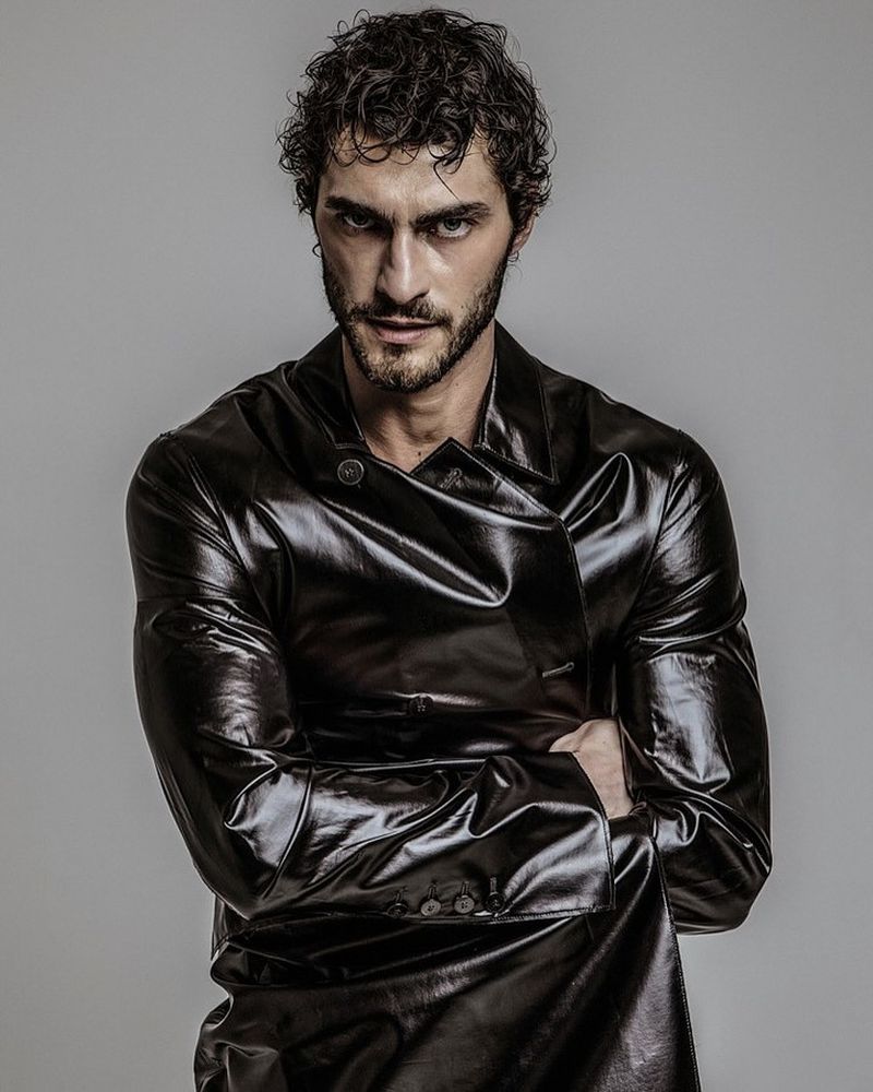 Portrait of a man with dark curly hair, beard, and a shiny black jacket, arms crossed in front of a plain grey studio backdrop.