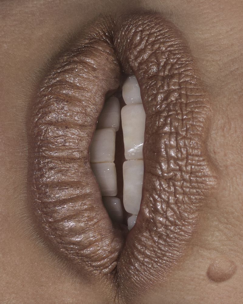 Extreme close-up of lips wearing metallic taupe lipstick, vertically framed and parted to reveal teeth.