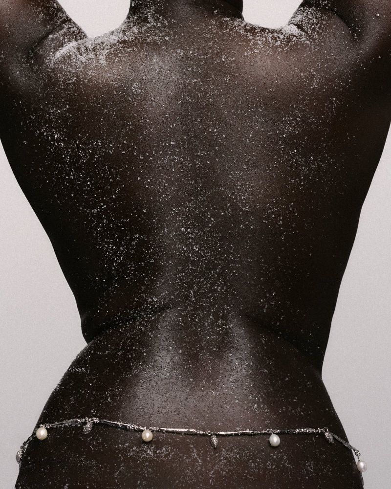 Close-up of a model's back dusted with fine white powder, wearing a delicate pearl and silver belly chain against a studio backdrop.