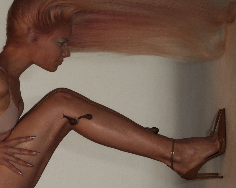 Blonde woman with long hair and high heels poses against a neutral backdrop, showcasing a minimal makeup look and intricate nail art.