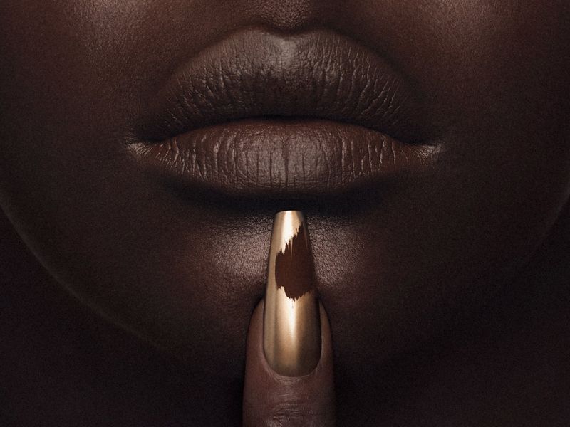 Macro close-up of a Black model's dark brown matte lips and a single finger with a metallic gold nail placed on the chin.