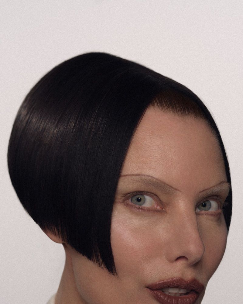 Close-up portrait of a model with sleek dark bob hair, lightened brows, and dark lipstick against a neutral backdrop.
