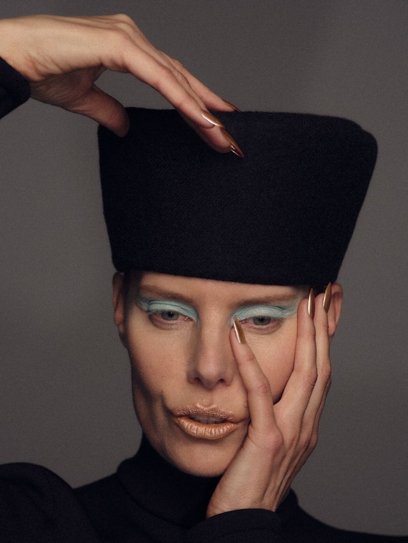 Model Hella Lund wearing a tall black felt hat with light blue eyeshadow and metallic nails against a grey backdrop.