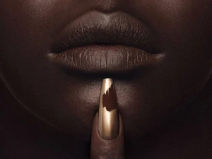 Macro close-up of a Black model's dark brown matte lips and a single finger with a metallic gold nail placed on the chin.