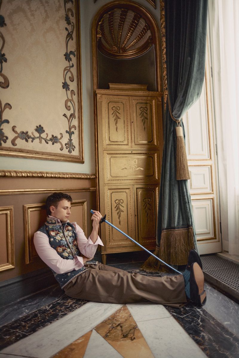 Man in an embroidered floral vest and brown trousers sitting on an ornate marble floor holding a fencing foil in an opulent room.