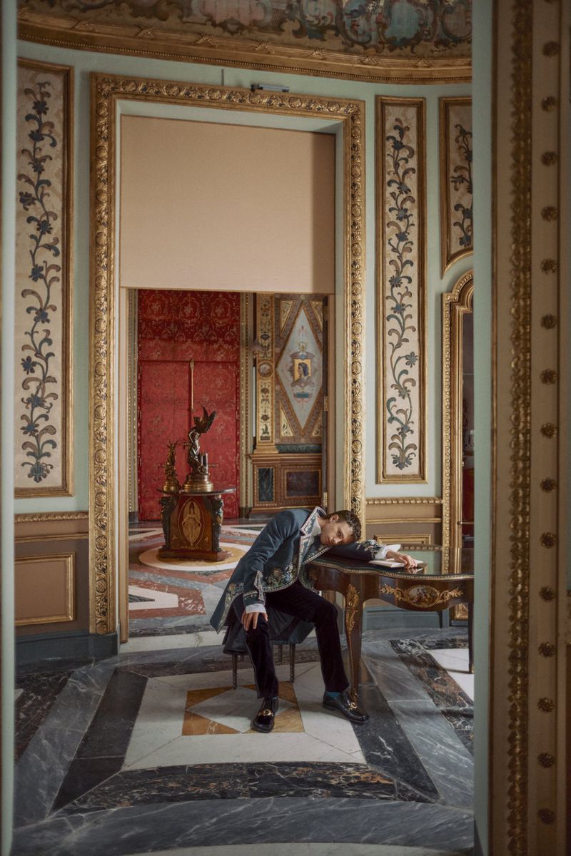 A man in an ornate embroidered coat rests his head on an antique desk inside an opulent gilded room with patterned marble floors.