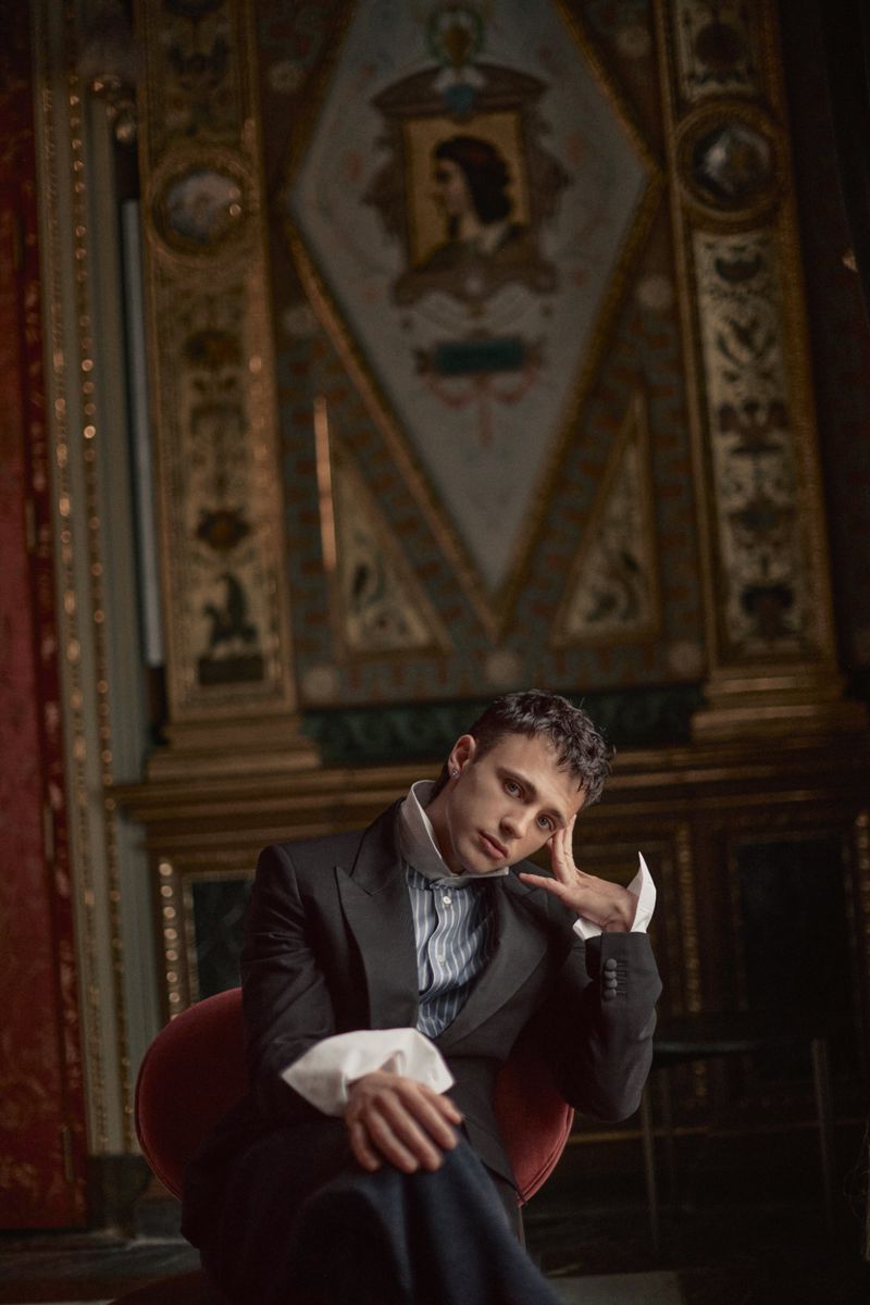 Man in a dark tailored suit with exaggerated white cuffs seated in front of an ornate classical wall panel.