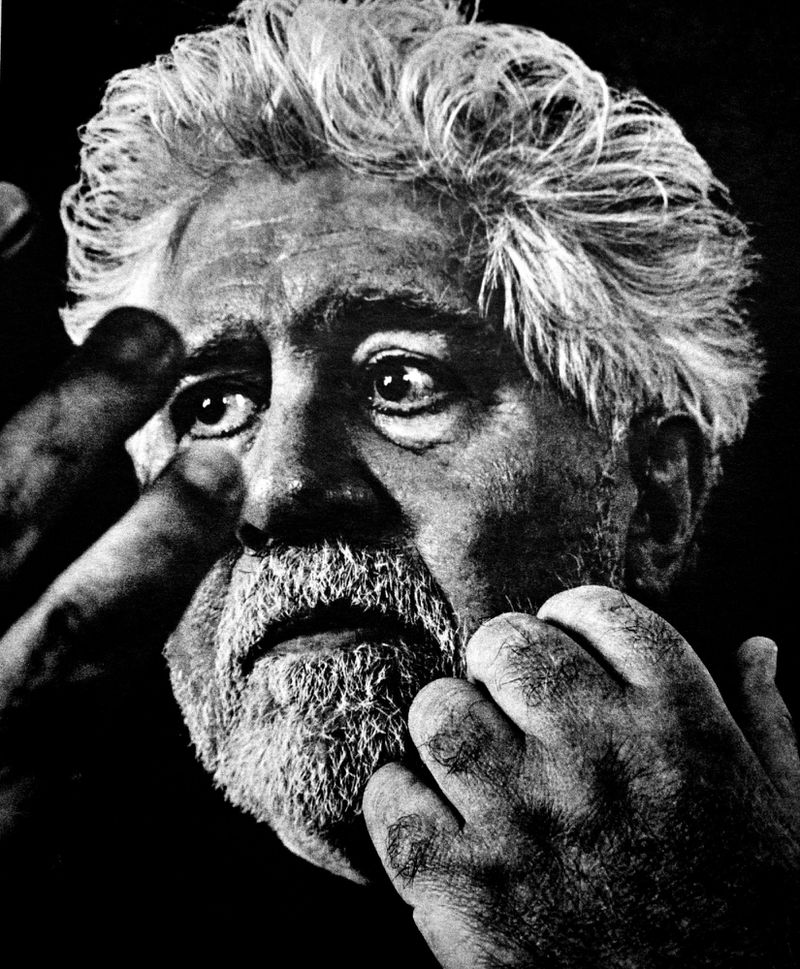 Monochrome close-up portrait of filmmaker Pedro Almodovar with hands framing his face against a dark background.