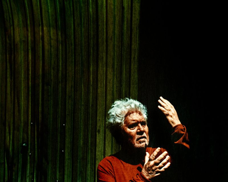 Pedro Almodóvar gesturing with both hands in dramatic low-key lighting beside a textured wooden wall.