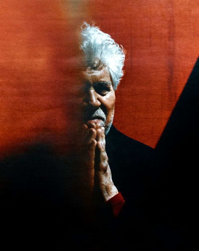 Portrait of filmmaker Pedro Almodóvar with hands clasped against a textured red background in warm chiaroscuro light.