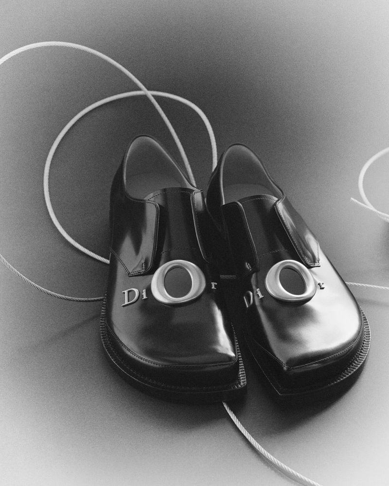 Pair of glossy black leather dress shoes with silver Dior lettering hardware, surrounded by a looping white cord.