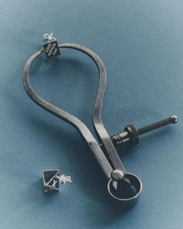 Vintage metal caliper holding a silver Louis Vuitton cube earring against a muted blue surface, with a second earring lying nearby.