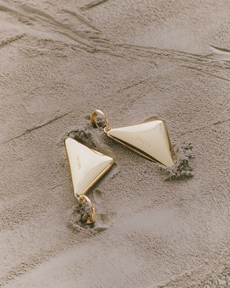 A pair of polished gold Prada triangle earrings resting partially submerged in fine textured grey sand.