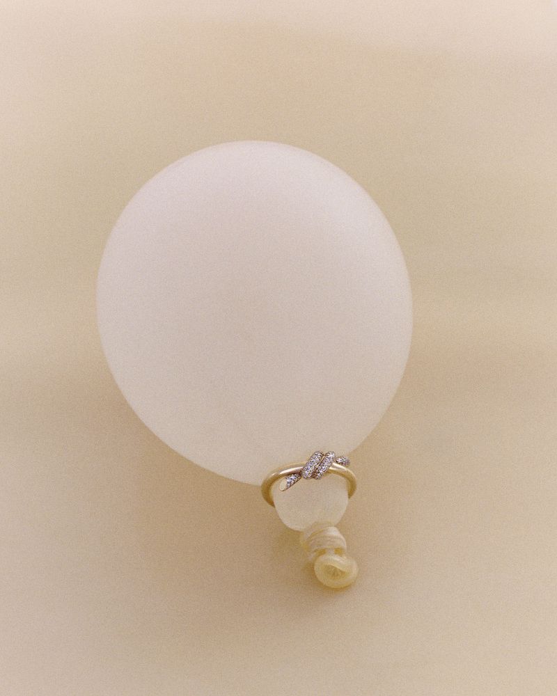 Close-up studio shot of a white balloon adorned with a gold and rhinestone ring, set against a soft, neutral-toned background.