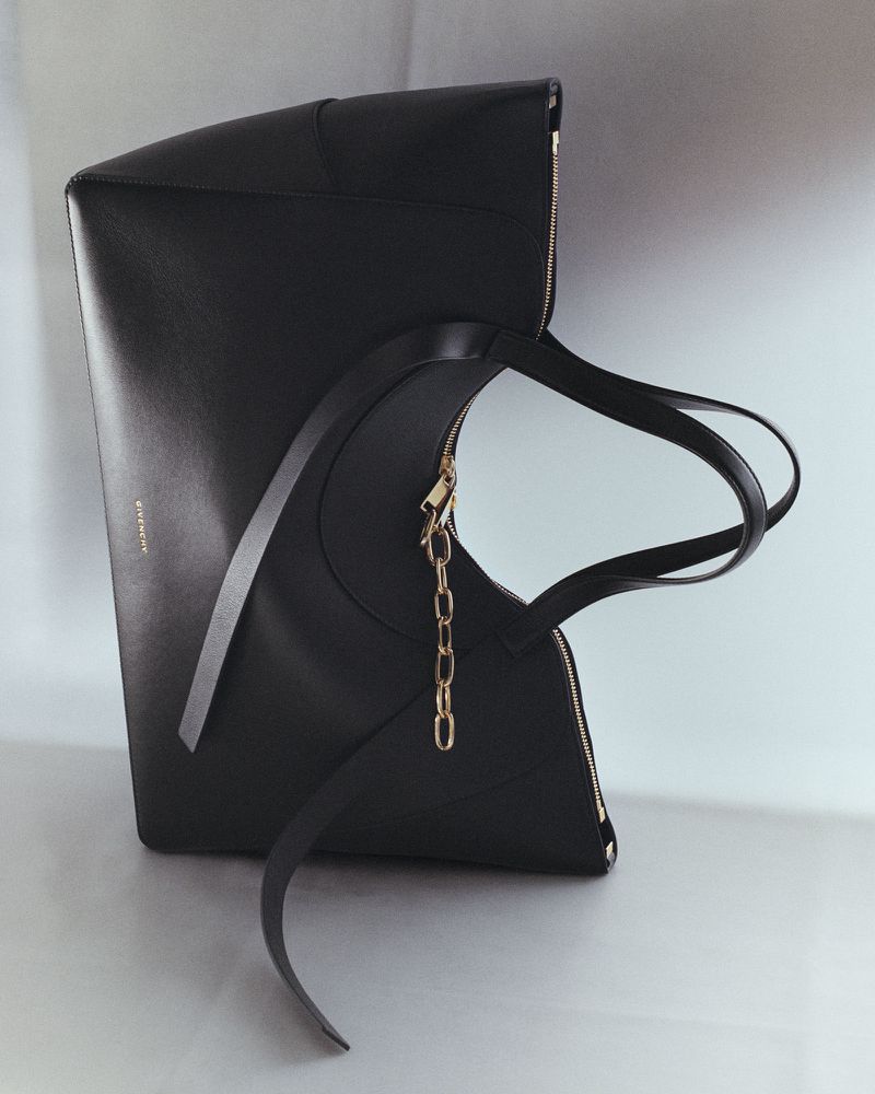 Sculptural black leather tote bag with a gold chain pull and curved handle against a soft grey studio background.
